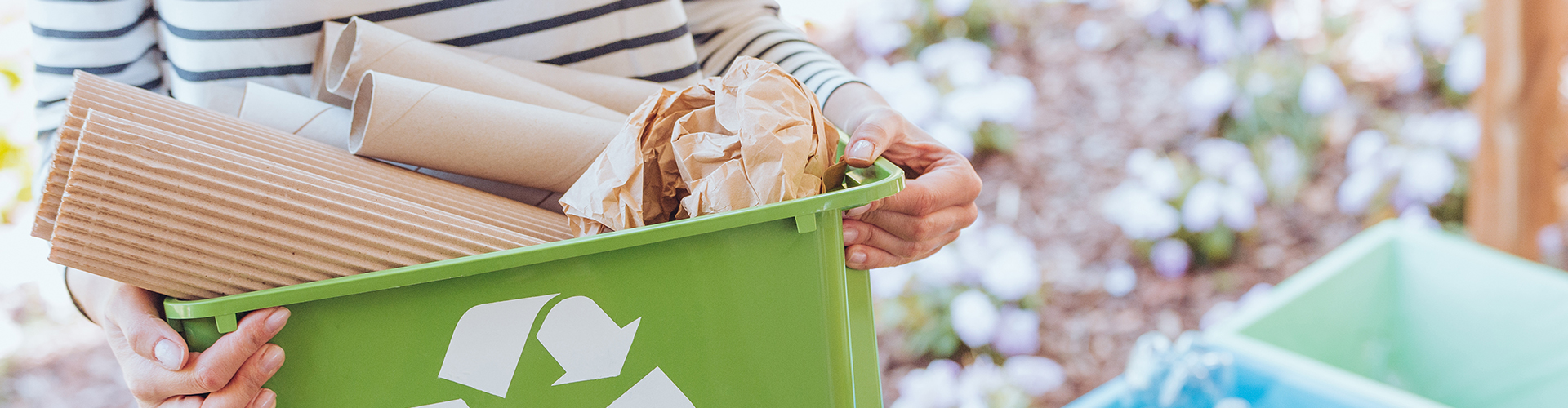Stockfoto zum Thema Müllentsorgung. Man sieht auf dem Foto eine Frau, die einen kleinen Behälter mit Recyclingsymbol hält, in dem sich Papierabfälle befinden. Im Hintergrund sind verschwommen weitere Müllbehälter zur Mülltrennung zu sehen.